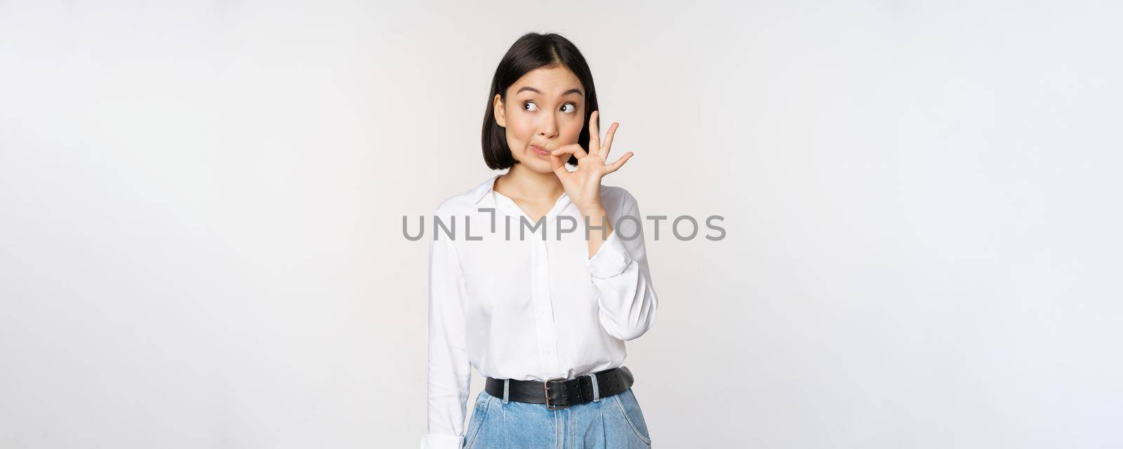 Mouth shut. Young asian woman seal lips, showing taboo dont speak, keep quiet sign, hiding secret, standing over white background.