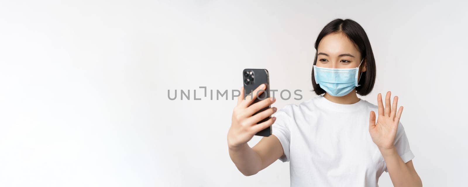 Health and covid-19 concept. Modern asian girl, student in medical mask, video chat with mobile phone, waving hand at smartphone app, standing over white background.