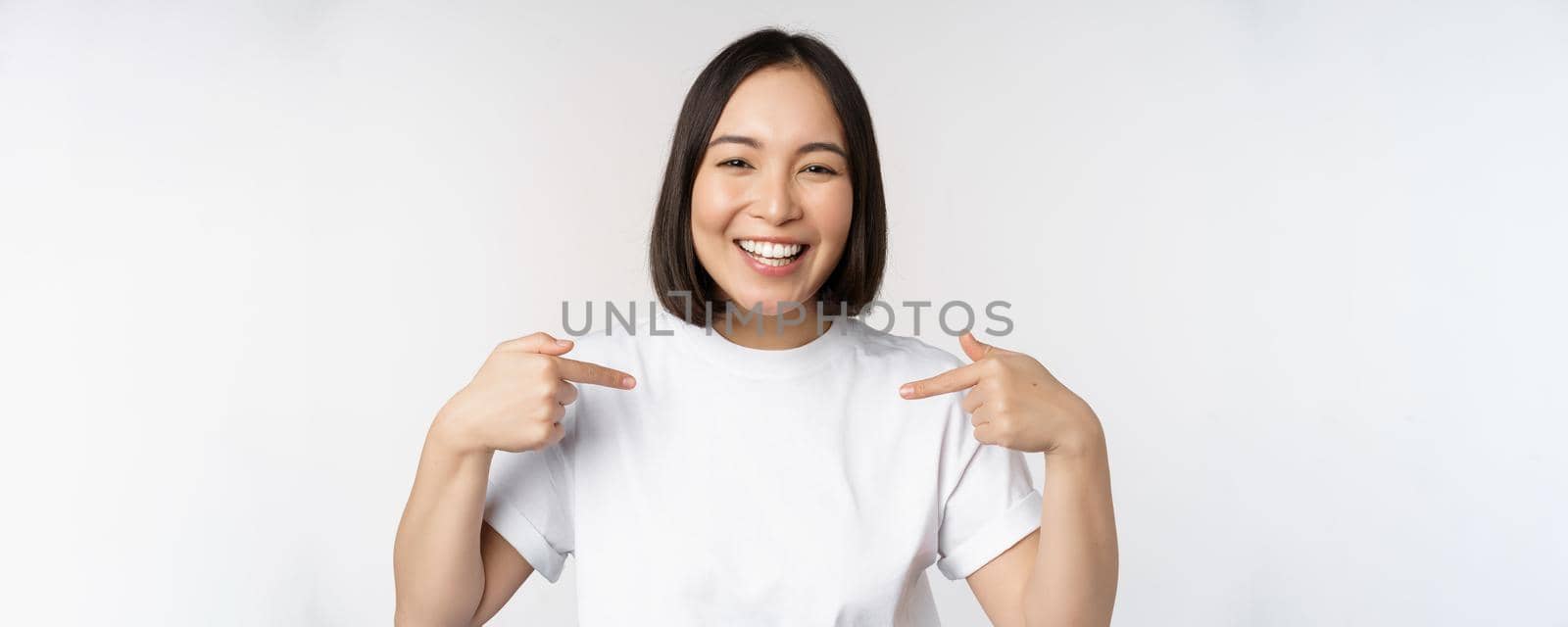 Happy and confident asian woman, student smiling and pointing at herself, self-promoting, showing logo on t-shirt, standing over white background.