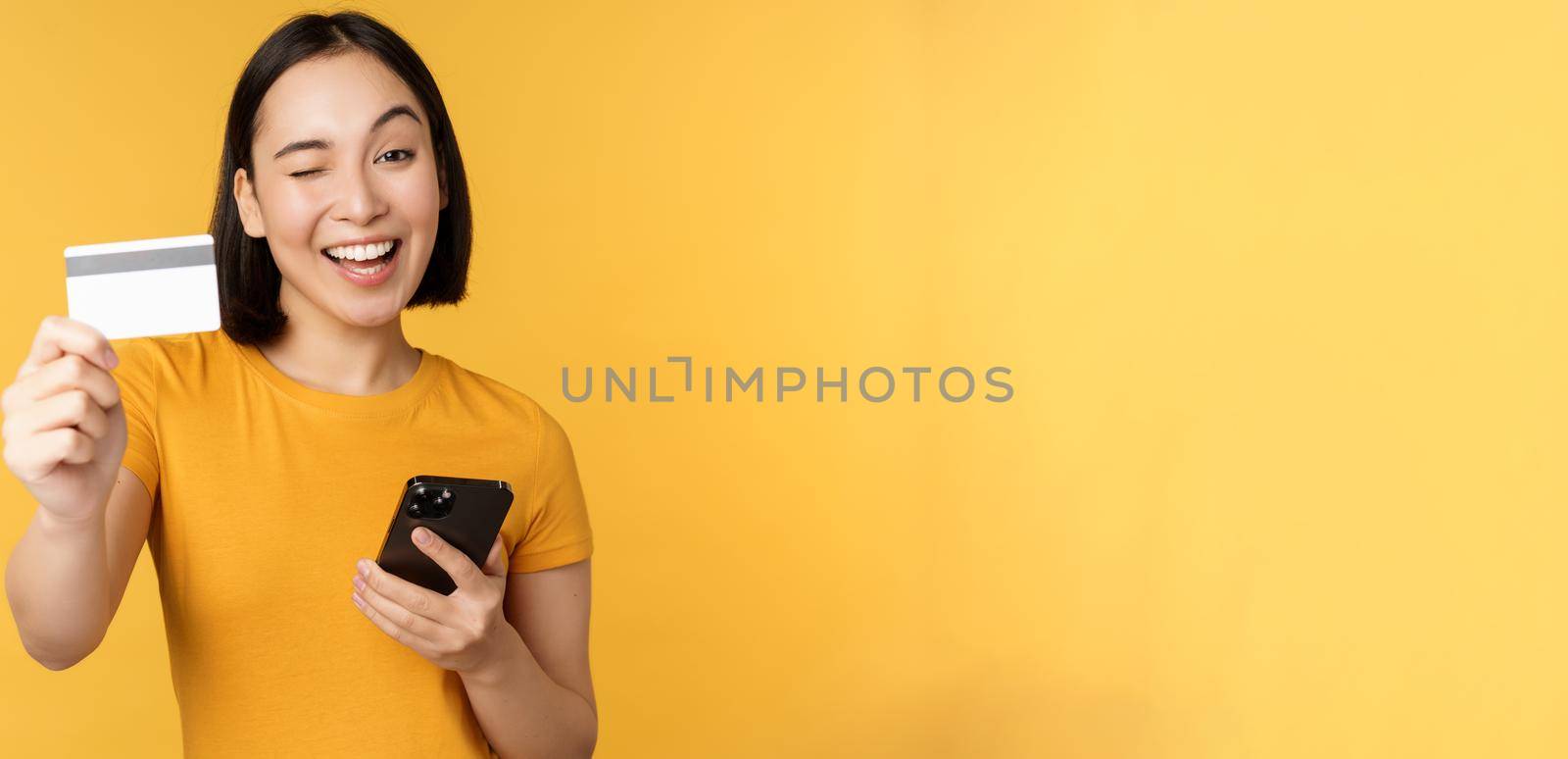 Joyful asian girl smiling, showing credit card and smartphone, recommending mobile phone banking, standing against yellow background.