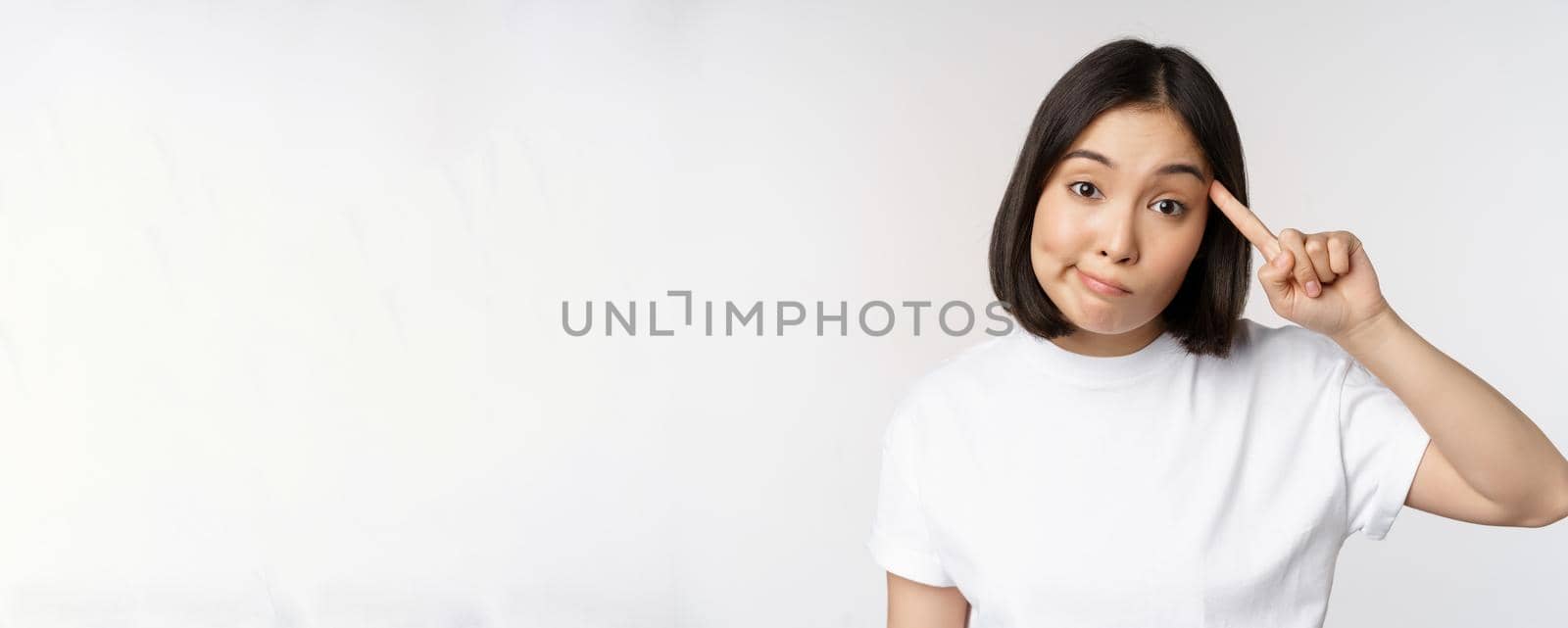 Are you crazy. Young asian woman roll finger on head, mocking someone stupid, standing in tshirt over white background. Copy space