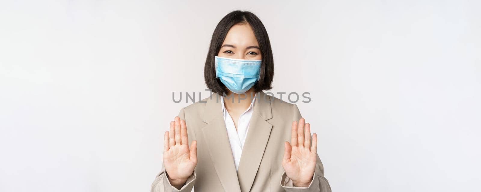 Coronavirus and work concept. Portrait of asian female office lady, woman at workplace wearing medical face mask and showing stop, prohibition gesture, white background.