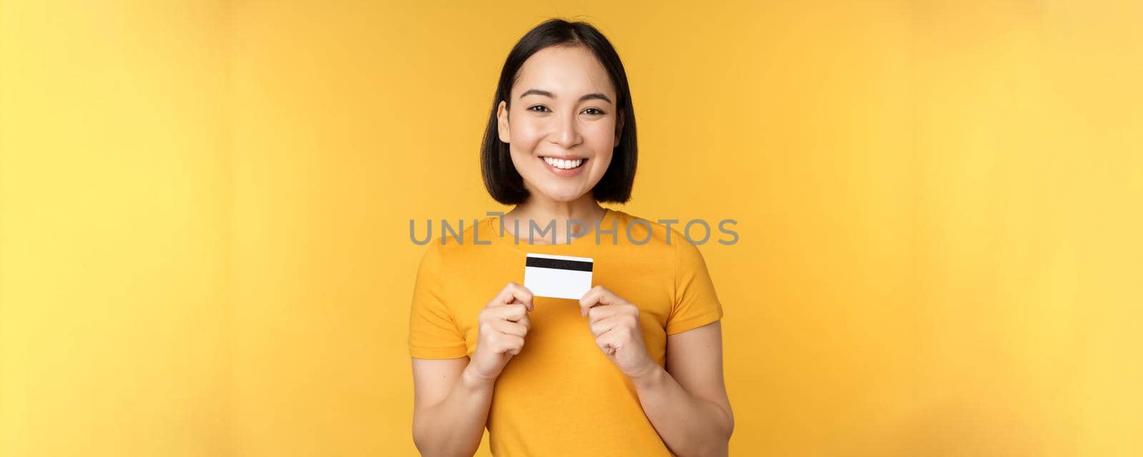 Beautiful asian woman showing credit card and smiling, recommending bank service, standing over yellow background. Copy space