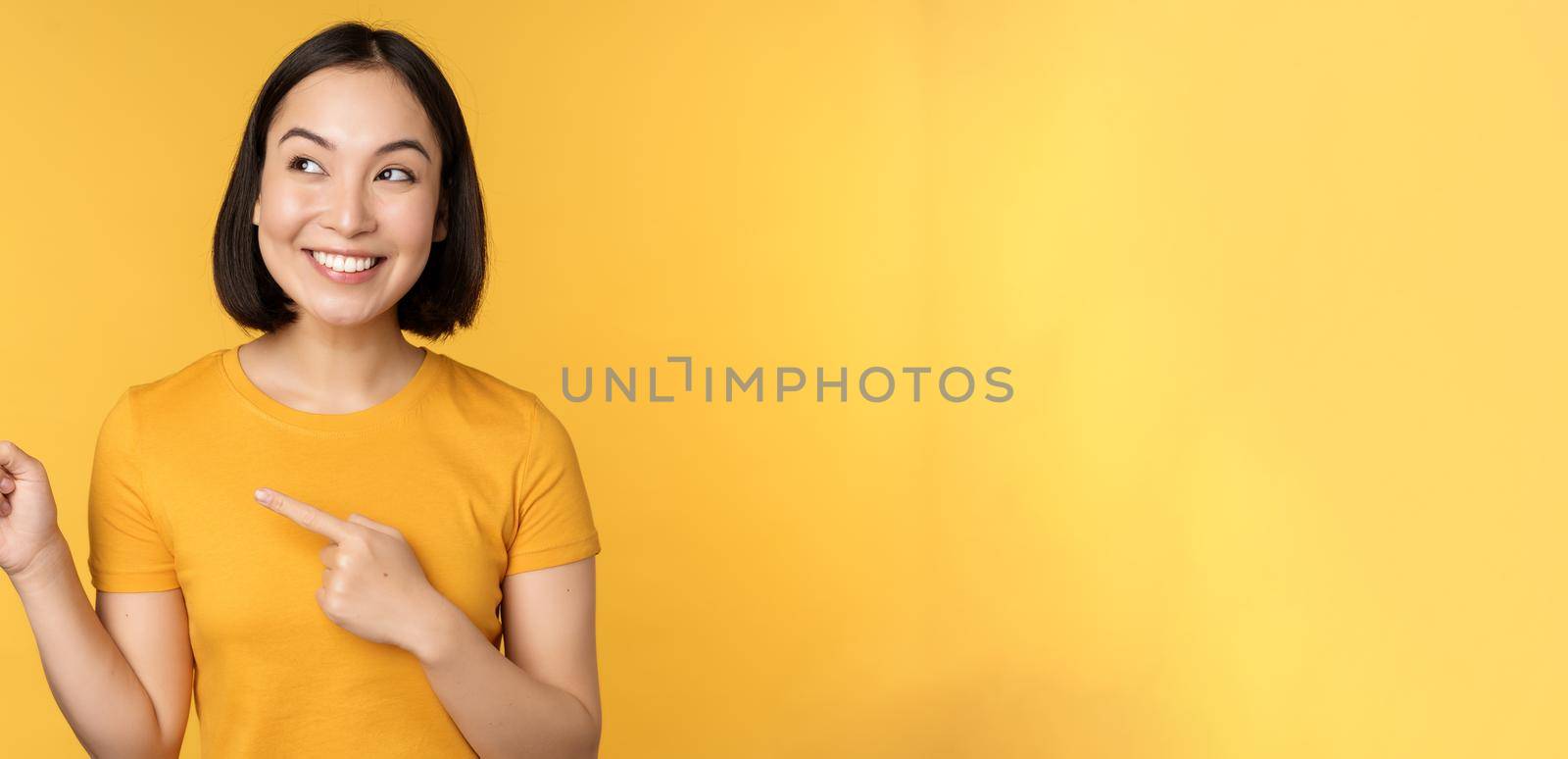 Portrait of happy asian girl pointing fingers and looking left, smiling amazed, checking out promo banner, showing advertisement against yellow background by Benzoix