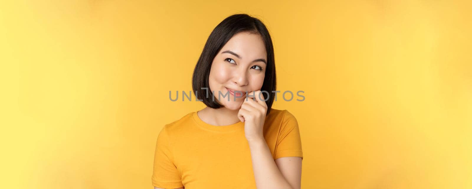 Close up portrait of cute asian girl smiling, thinking, looking up thoughtful, standing in tshirt over yellow background by Benzoix
