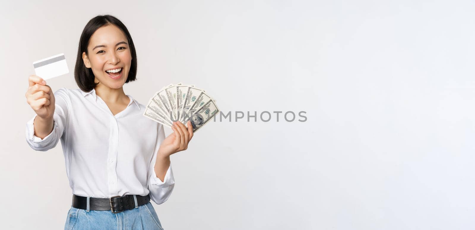 Happy korean woman holding credit card and money dollars, smiling and laughing, posing against white studio background.