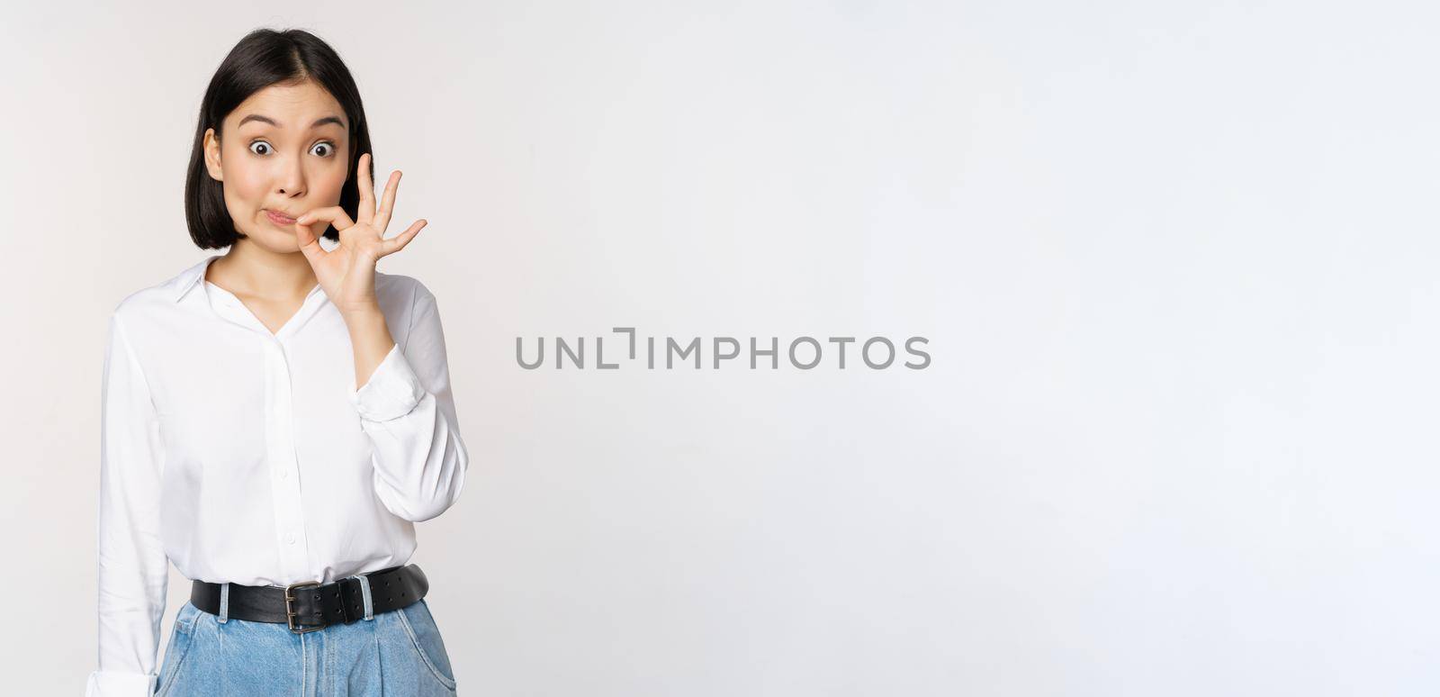 Mouth shut. Young asian woman seal lips, showing taboo dont speak, keep quiet sign, hiding secret, standing over white background.