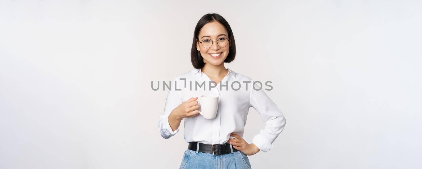 Happy young energetic asian woman smiling, drinking, holding cup mug of coffee, standing confident against white background.