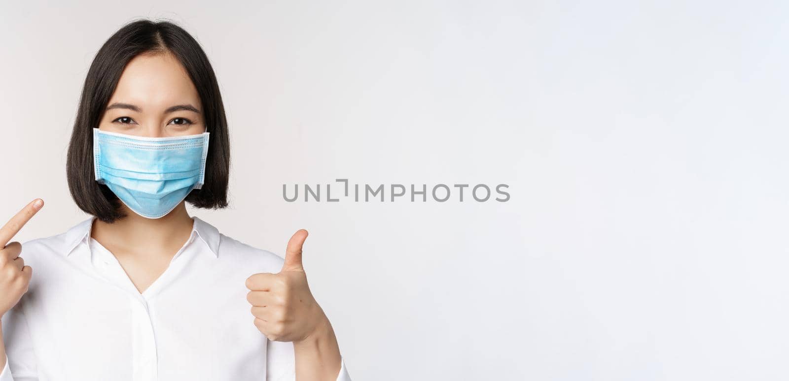 Portrait of asian girl in medical mask showing thumbs up sign and pointing at her covid protection, standing over white background.