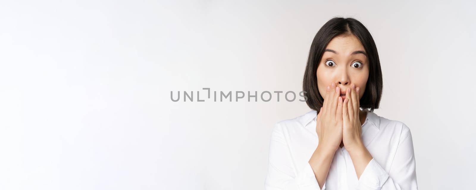 Close up face of asian woman gasping, looking shocked and speechless, holding hands near mouth and staring at camera impressed, white background.