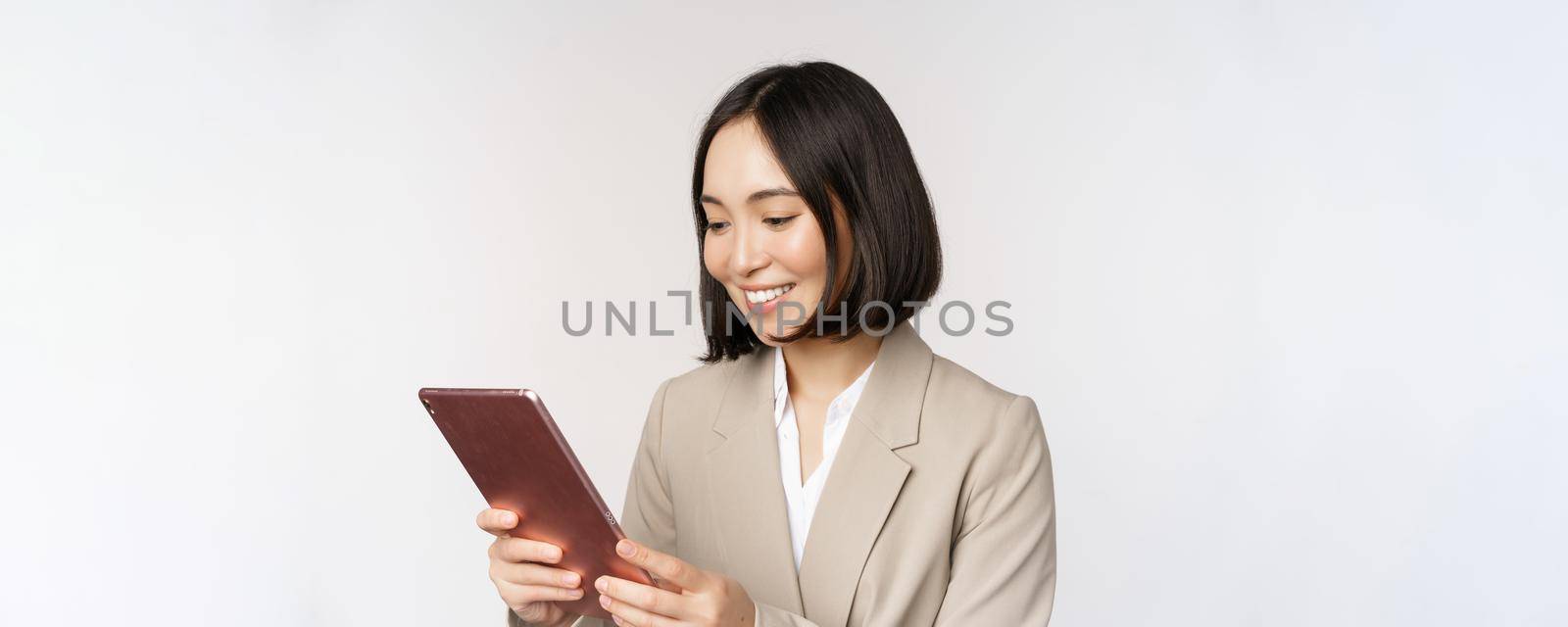 Image of asian businesswoman using digital tablet, looking at gadget and smiling, working, standing against white background.