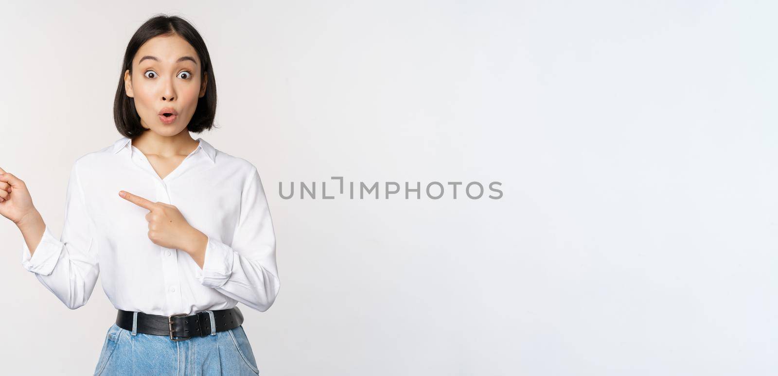 Portrait of excited young asian woman, office lady pointing fingers left at discount, showing sale banner, standing over white background by Benzoix