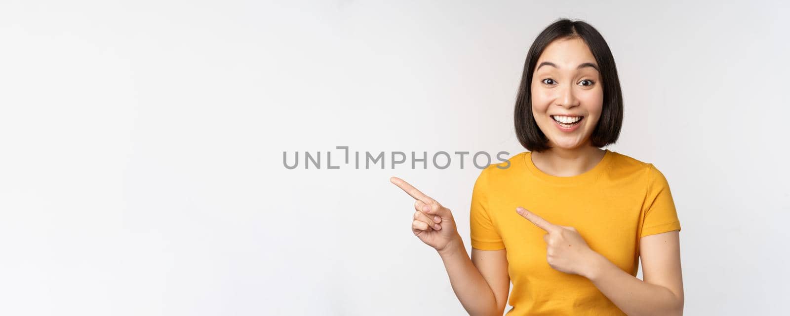 Portrait of smiling asian brunette girl in yellow tshirt, pointing fingers left, showing copy space, promo deal, demonstrating banner, standing over white background.