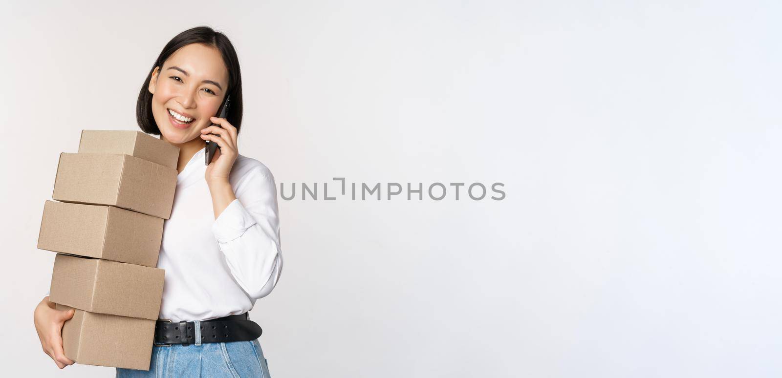Image of young asian businesswoman answer phone call while carrying boxes for delivery, posing against white background.