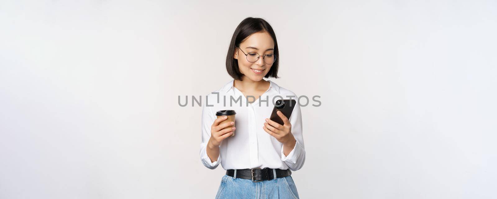 Image of modern asian woman looking at mobile phone, drinking takeaway coffee, wearing glasses, using smartphone app, standing over white background.