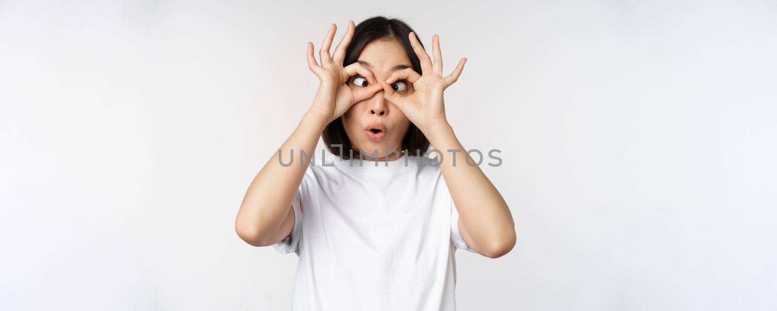 Funny young asian woman, korean girl making eyes glasses gesture, looking happy at camera, standing over white background by Benzoix