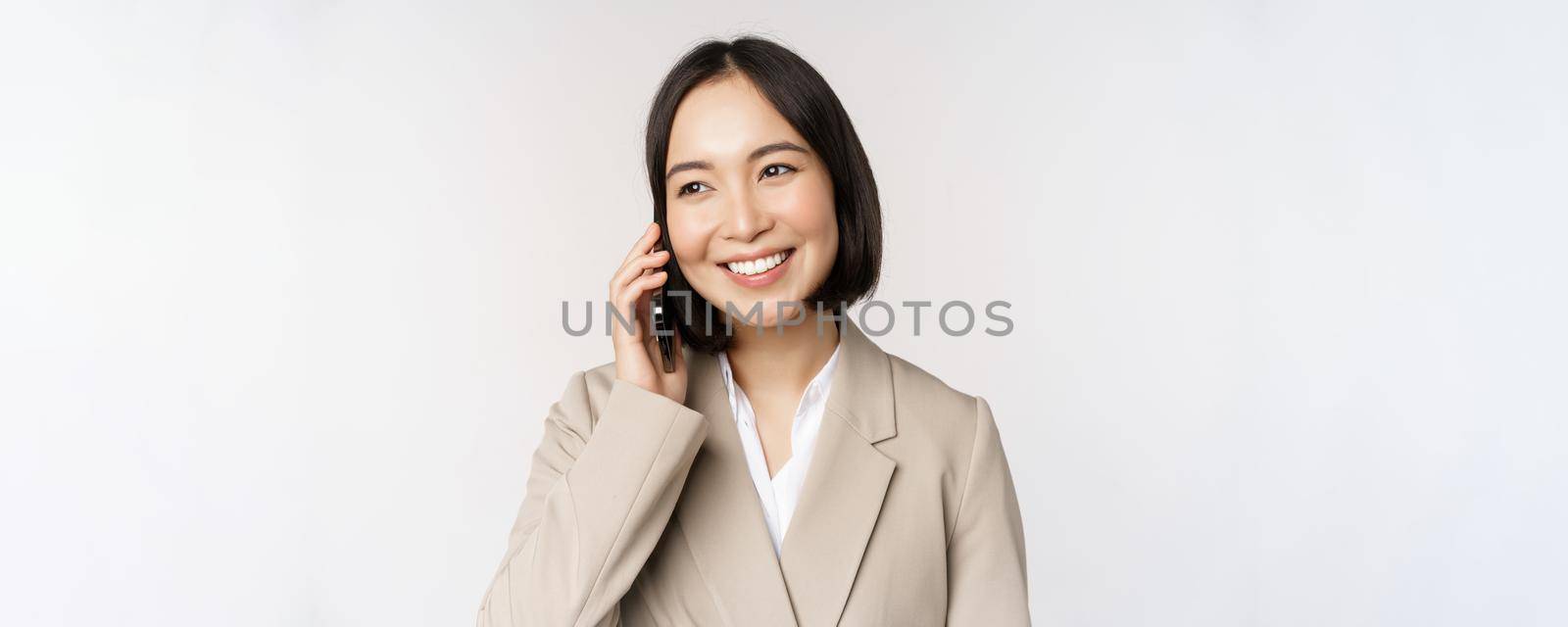 Smiling corporate woman in suit, talking on mobile phone, having a business call on smartphone, standing over white background.