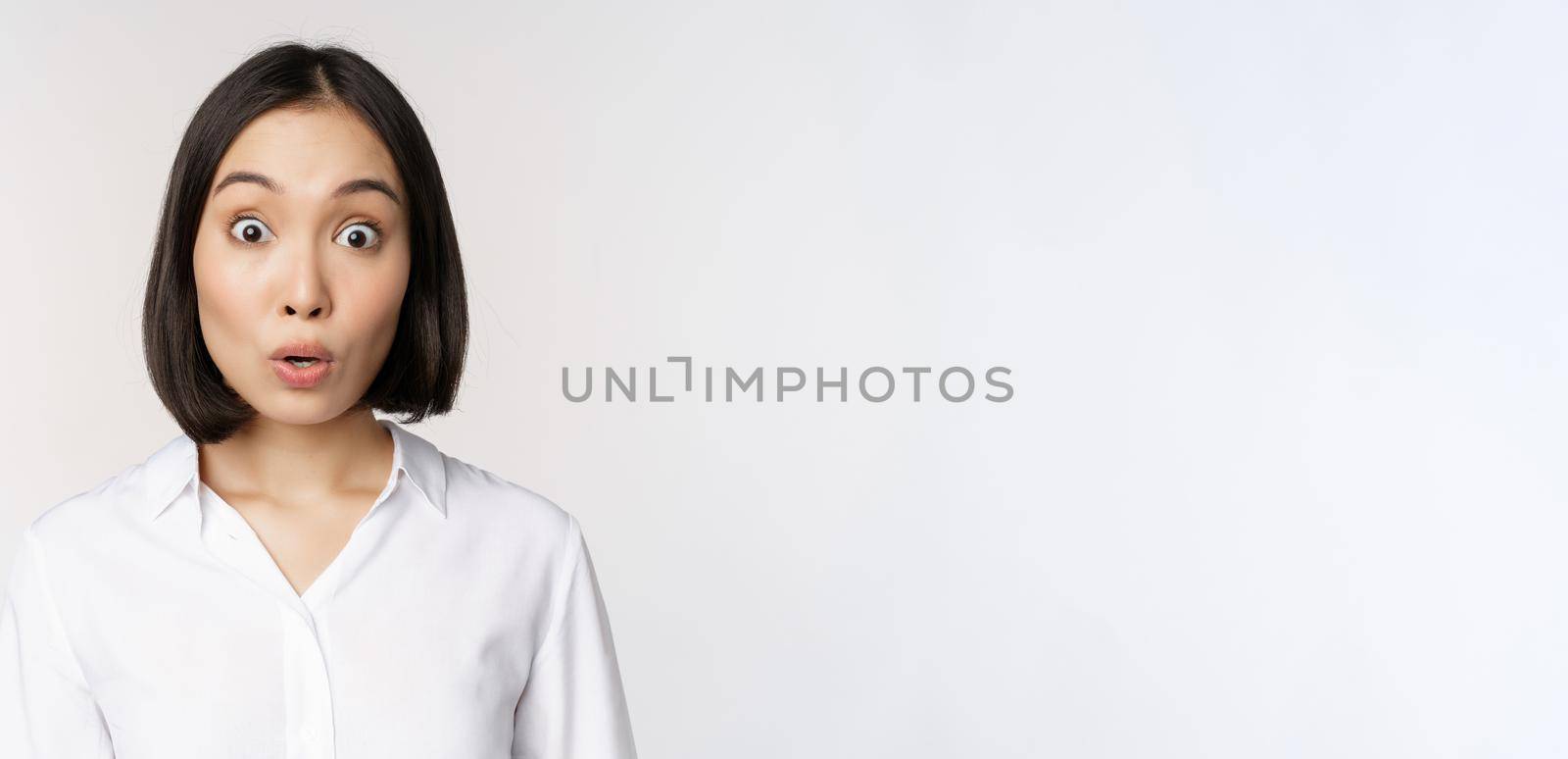 Close up portrait of young asian female model looking amazed at camera, smiling white teeth, standing against white background.