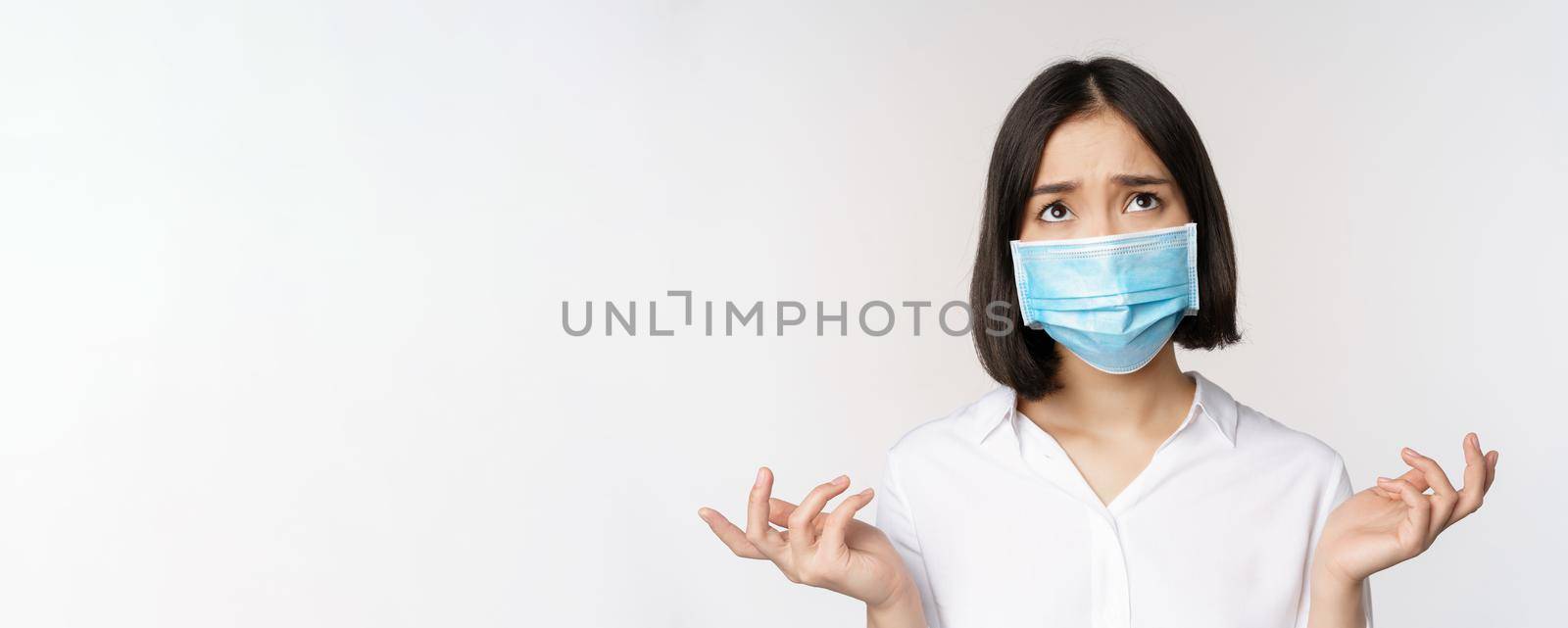 Distressed and miserable young asian woman in face mask, looking up, looking up sad, standing over white background.