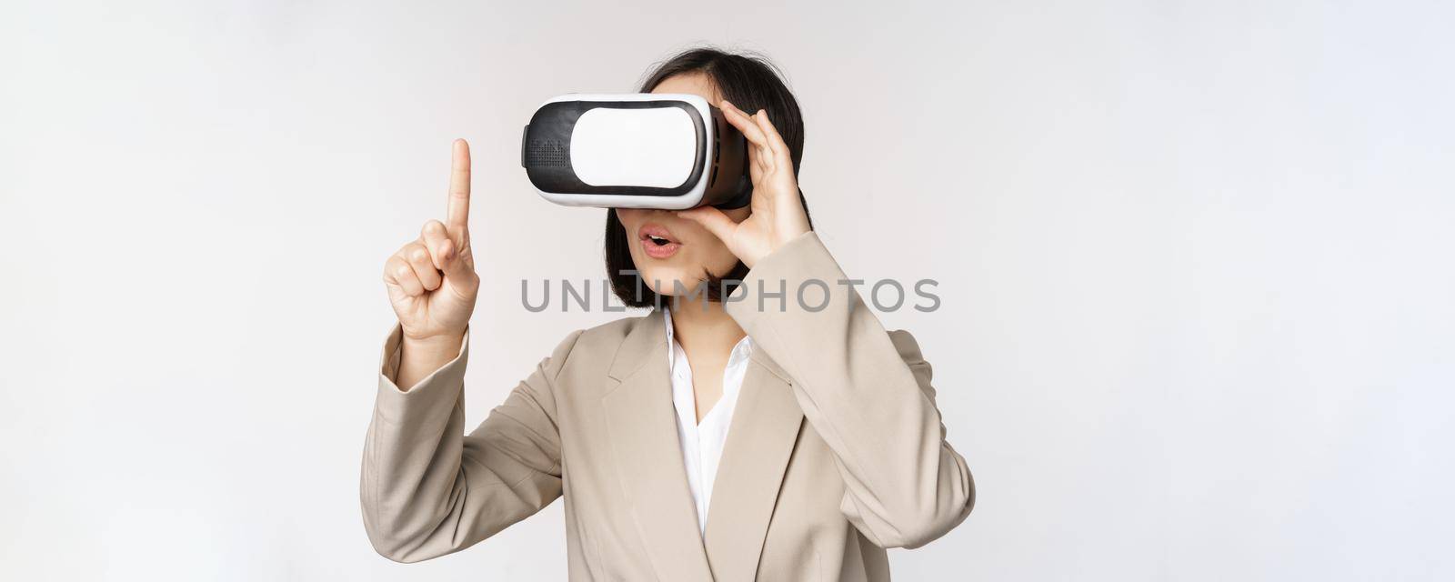 Amazed business woman in suit using virtual reality glasses, looking amazed in vr headset, standing over white background.