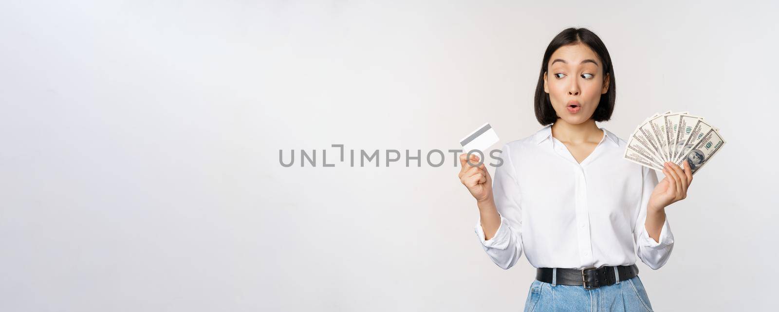 Excited korean girl looking at credit card, holding money cash, posing against studio background by Benzoix