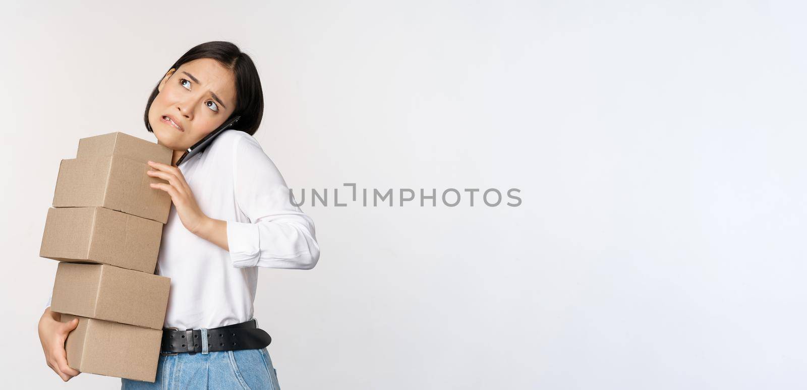 Young asian businesswoman answer phone call, talking on mobile while carrying pile of boxes with orders, standing over white background by Benzoix