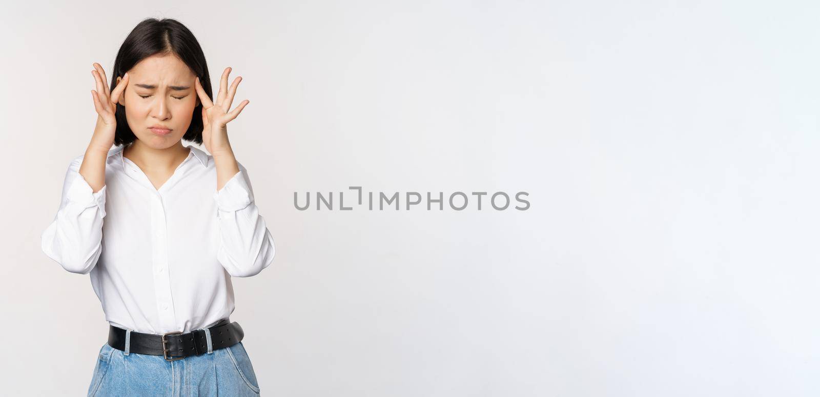 Image of distressed young asian businesswoman, female lady touching head, rubbing temples, has migraine, standing over white background.