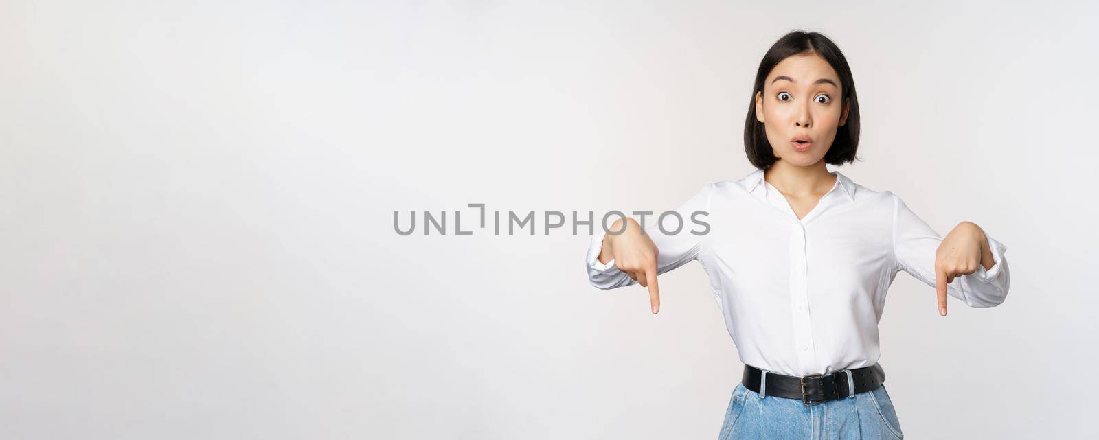 Portrait of young asian woman pointing fingers down and smiling, showing banner, click on link below gesture, inviting people to follow, standing over white background.