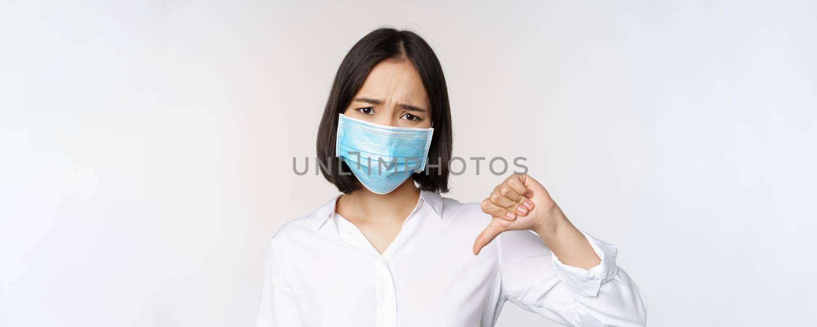 Portrait of asian woman in medical face mask showing thumbs down with disappointed, tired face expression, standing over white background.