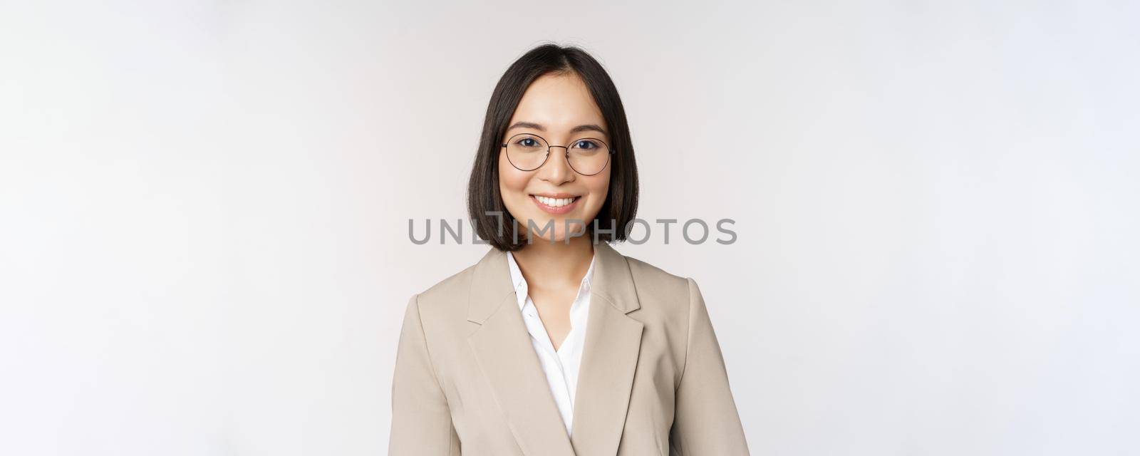 Portrait of young asian saleswoman in glasses, wearing beige suit, smiling and looking confident at camera, white background.