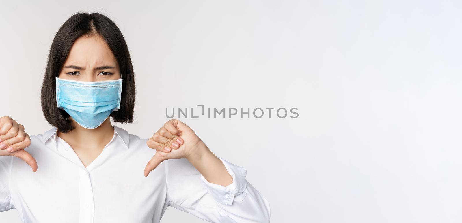 Portrait of asian woman in medical face mask showing thumbs down with disappointed, tired face expression, standing over white background by Benzoix