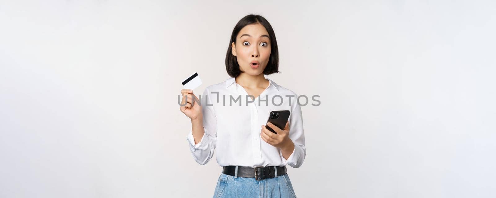 Online shopping concept. Image of surprised asian girl, holding credit card and smartphone, looking amazed in disbelief at camera, white background.