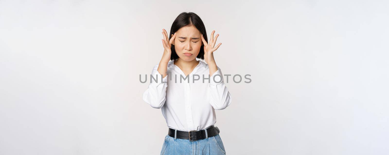 Image of distressed young asian businesswoman, female lady touching head, rubbing temples, has migraine, standing over white background.