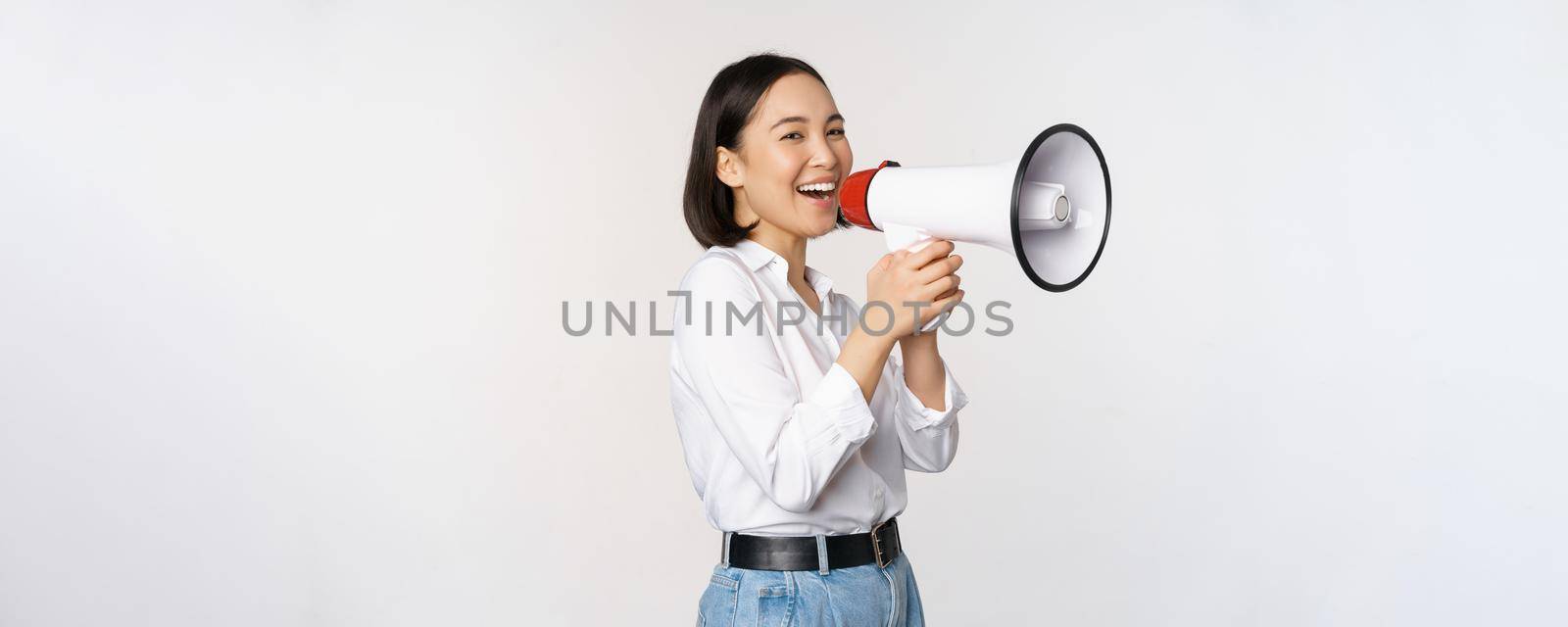 Image of young woman, korean activist, recruiter screaming in megaphone, searching, shouting at loudspeaker, standing over white background by Benzoix