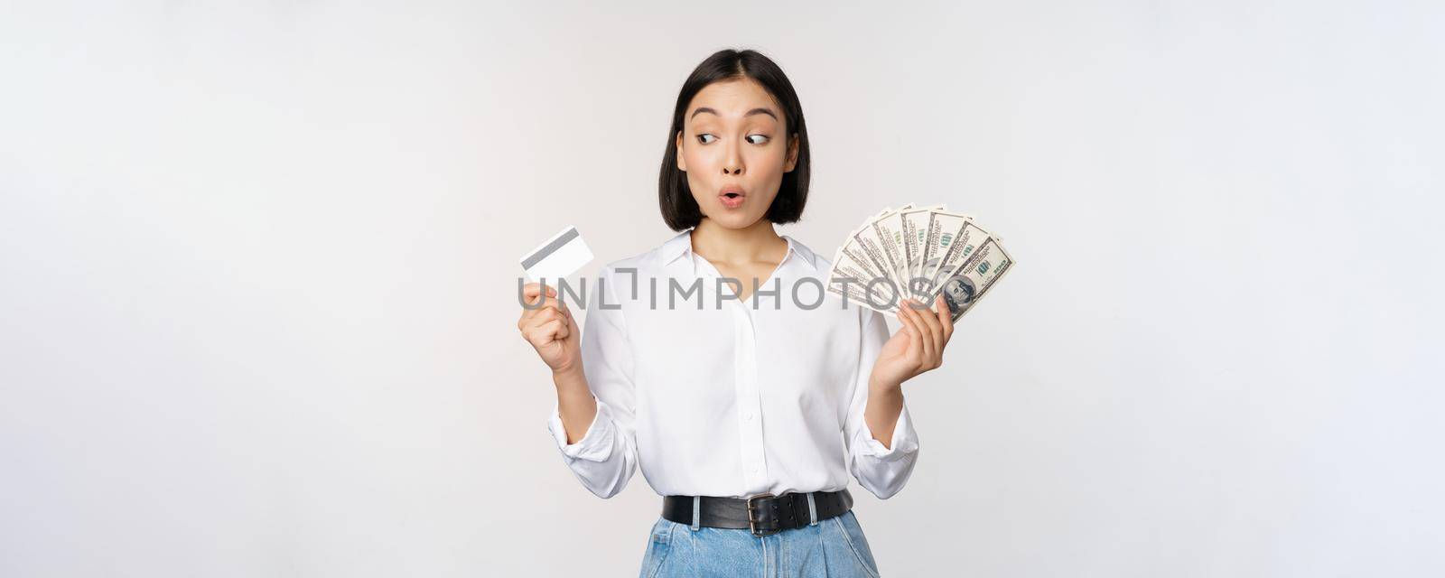 Excited korean girl looking at credit card, holding money cash, posing against studio background. Finance concept