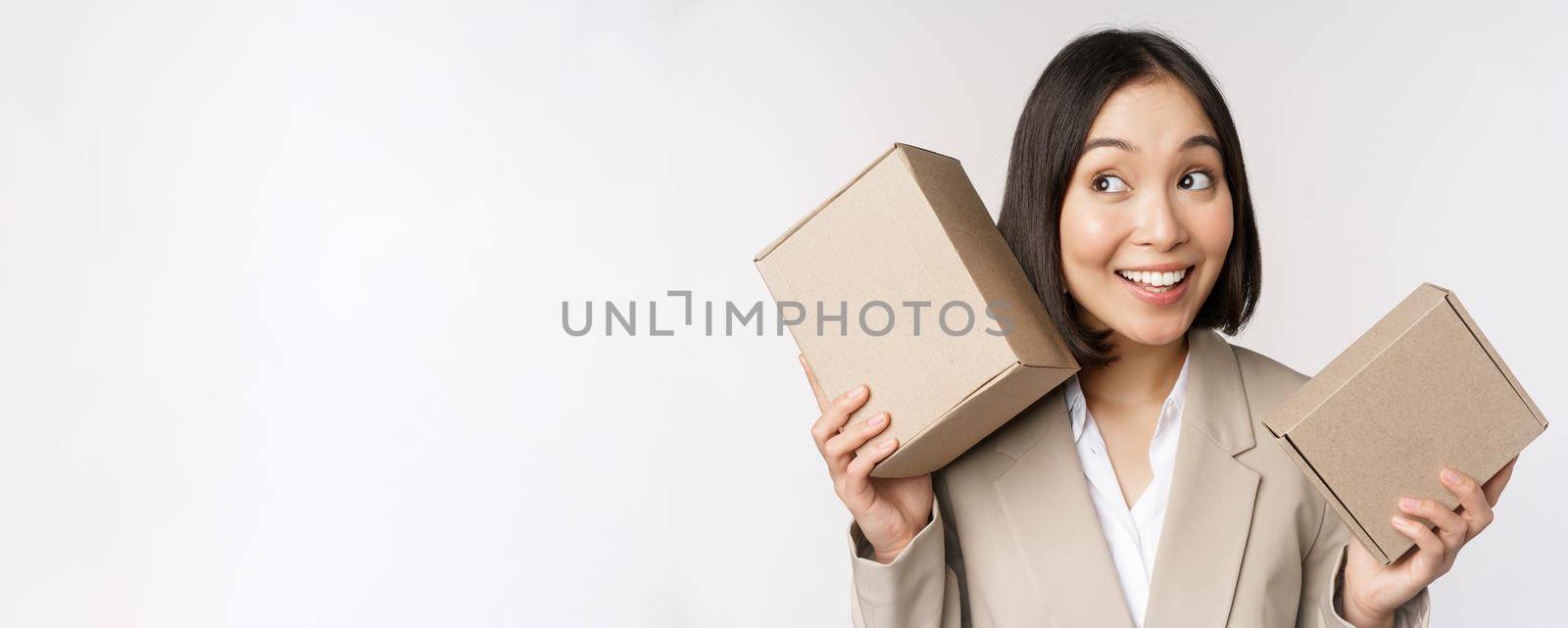 Image of asian saleswoman shaking boxes and guessing whats inside, smiling thoughtful, standing over white background. Copy space