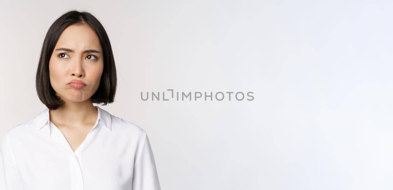 Close up head portrait of young asian woman looking upset and disappointed at left copy space, grimacing and frowning displeased, white background.