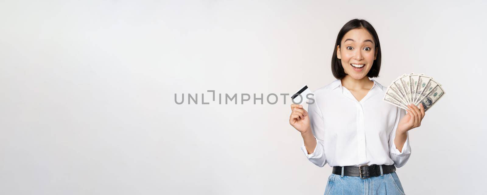 Portrait of enthusiastic asian woman holding money in cash and credit card, smiling amazed at camera, white background.