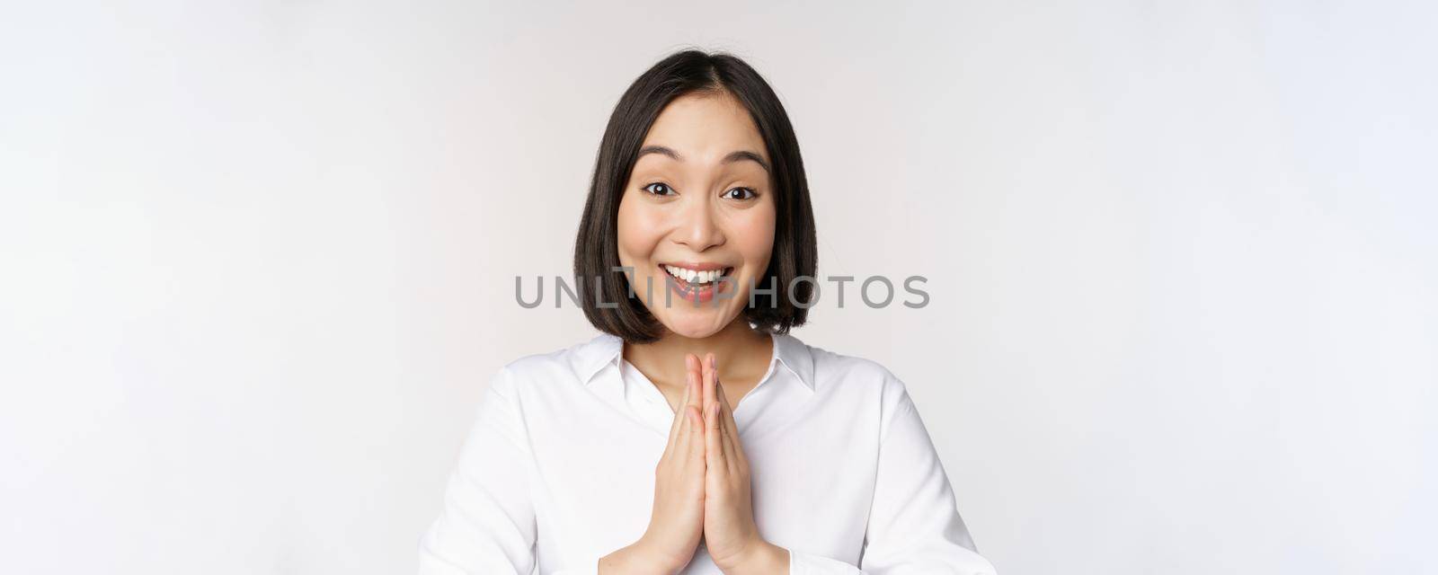 Close up portrait of young japanese woman showing namaste, thank you arigatou gesture, standing over white background by Benzoix