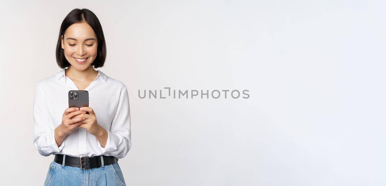 Stylish modern asian girl using mobile phone application, chatting on cellphone and smiling, standing in white blouse against studio background by Benzoix