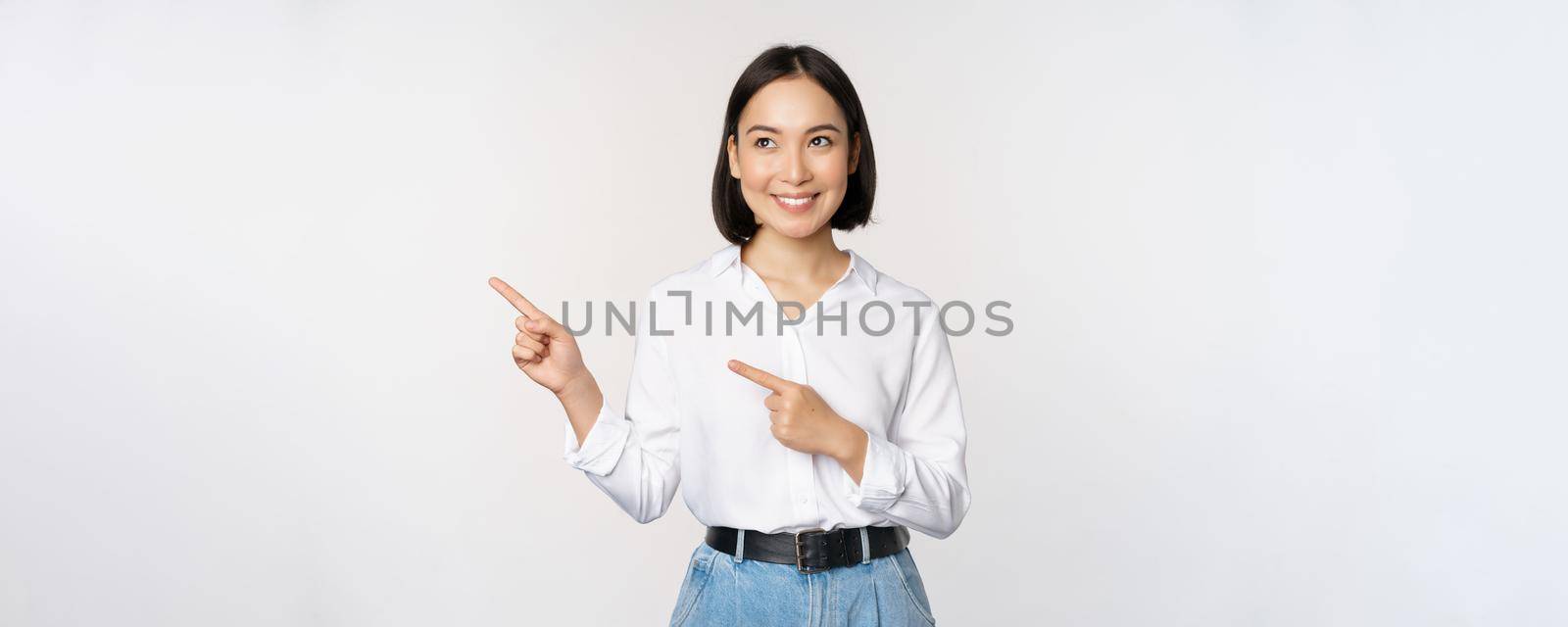 Portrait of attractive adult asian woman pointing, looking left with pleased smile, showing banner or logo aside, standing against white studio background by Benzoix
