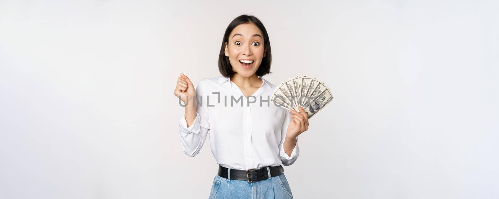 Enthusiastic young asian woman looking excited at camera, holding money dollars in hand, standing over white background by Benzoix