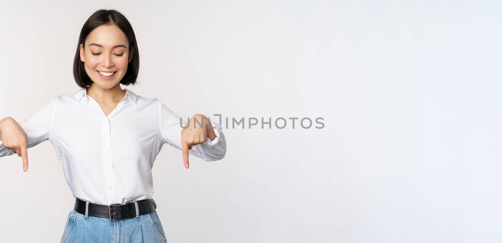 Portrait of happy asian woman pointing fingers down and looking below at advertisement, showing info banner, advertising, standing over white background by Benzoix