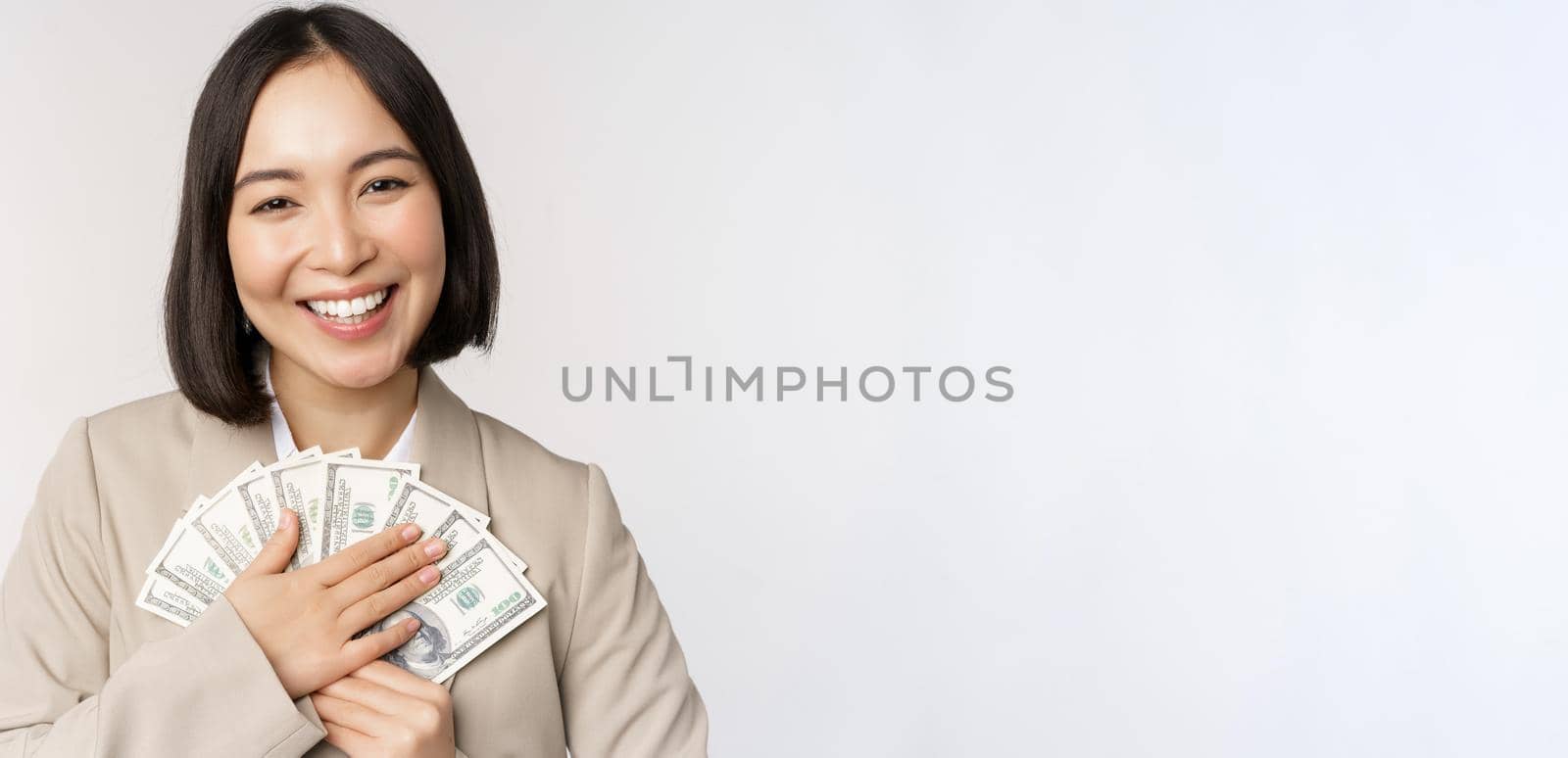 Happy asian businesswoman holding cash, hugging dollars money and smiling, standing over white background in suit by Benzoix