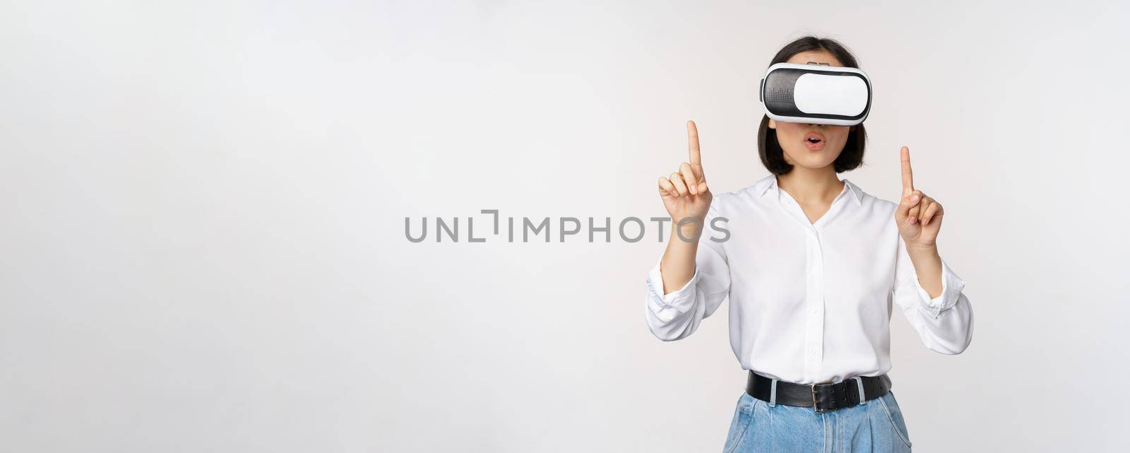 Image of asian woman in vr glasses pointing fingers up, looking amazed and surprised. Girl using virtual reality headset, concept of future and communication, white background.