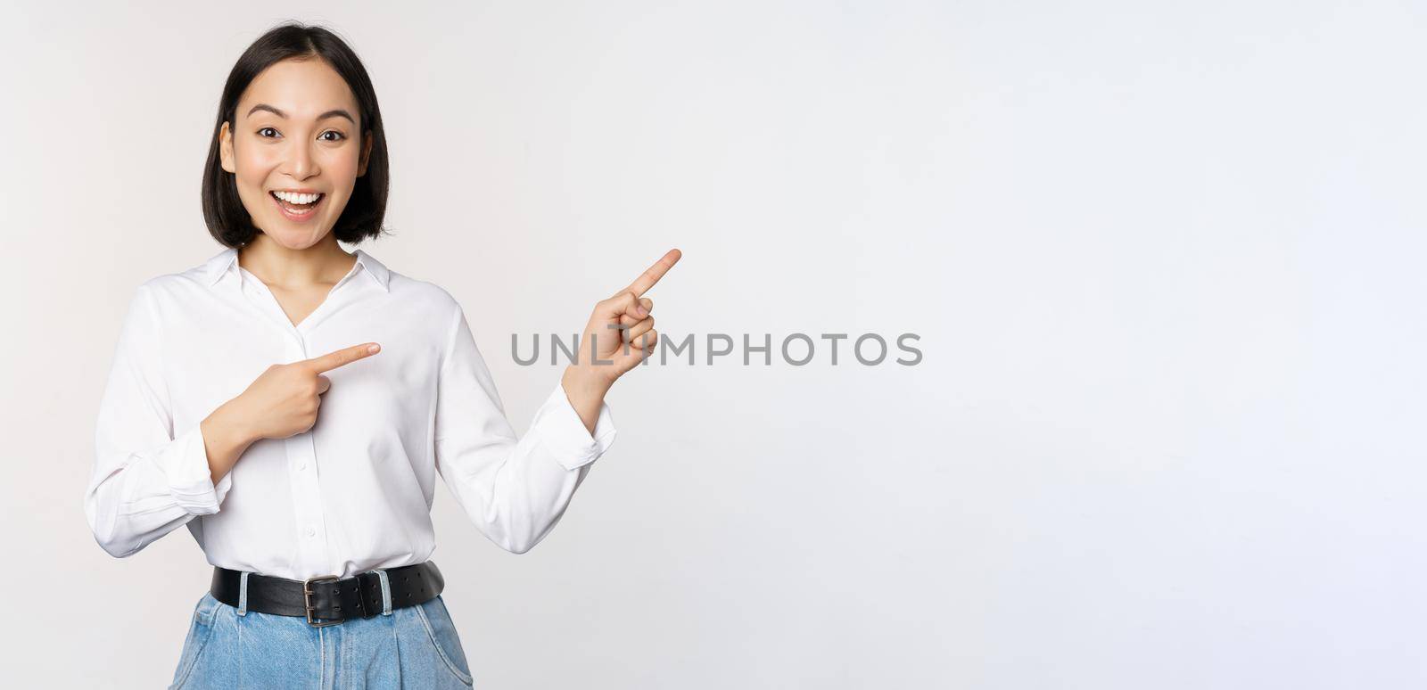 Portrait of beautiful asian woman, saleswoman pointing fingers right and showing info, banner or logo, sale advertisement, standing over white background.