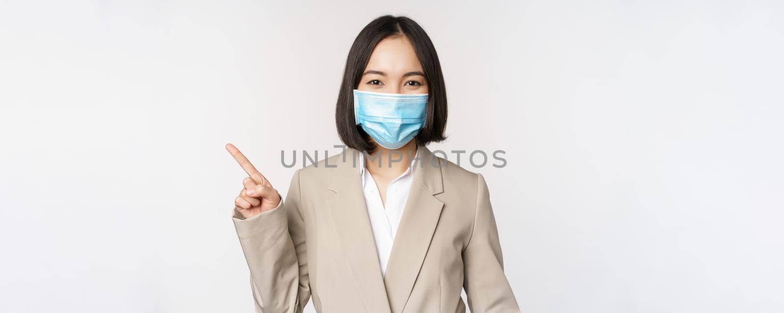 Coronavirus and work concept. Portrait of woman in medical face mask, pointing finger left, showing logo or banner, advertisement, white background.