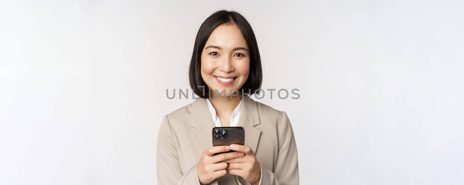 Image of asian businesswoman in suit, holding mobile phone, using smartphone app, smiling at camera, white background.