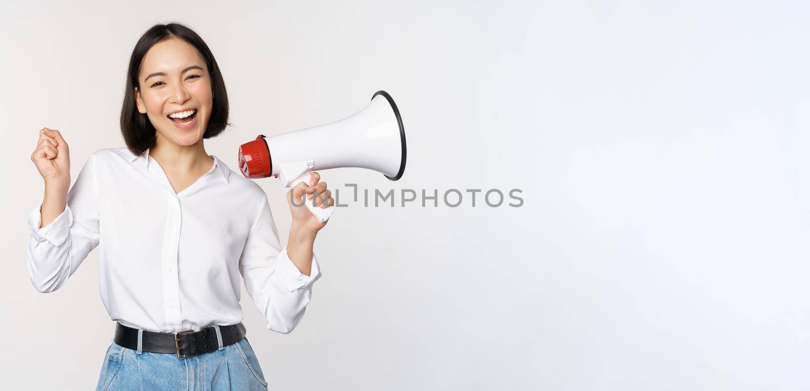 Smiling young asian woman posing with megaphone, concept of news, announcement and information, standing over white background.