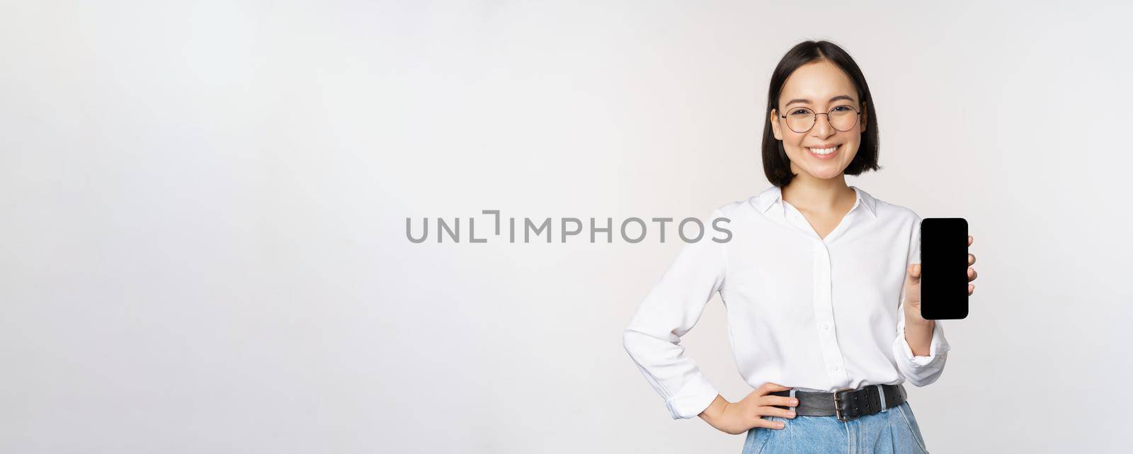 Smiling korean woman showing smartphone screen, demonstrating mobile application, standing over white background by Benzoix